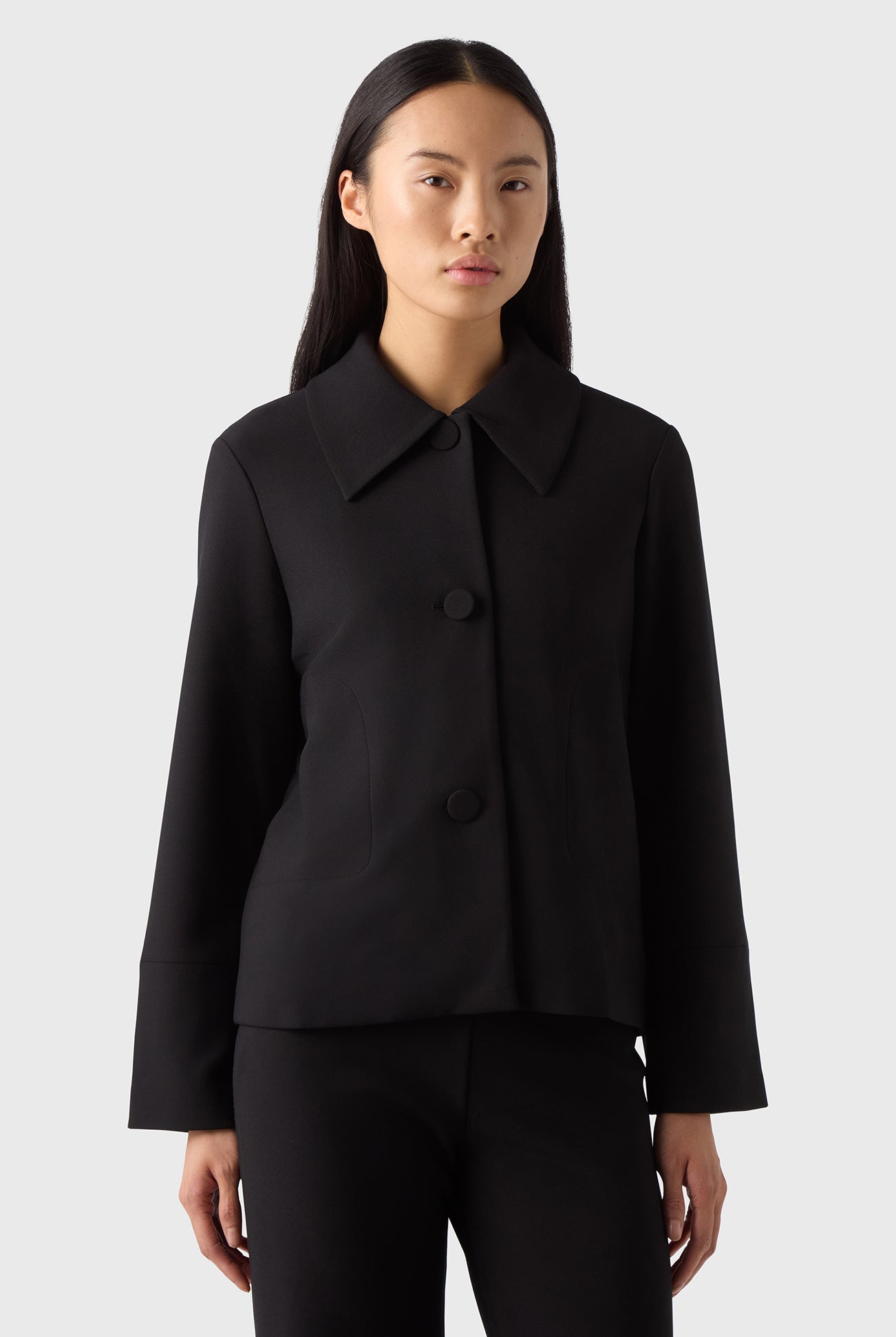 A woman standing against a white background wearing a navy blazer with a classic collar and tailored cut, paired with blue jeans and beige sandals.